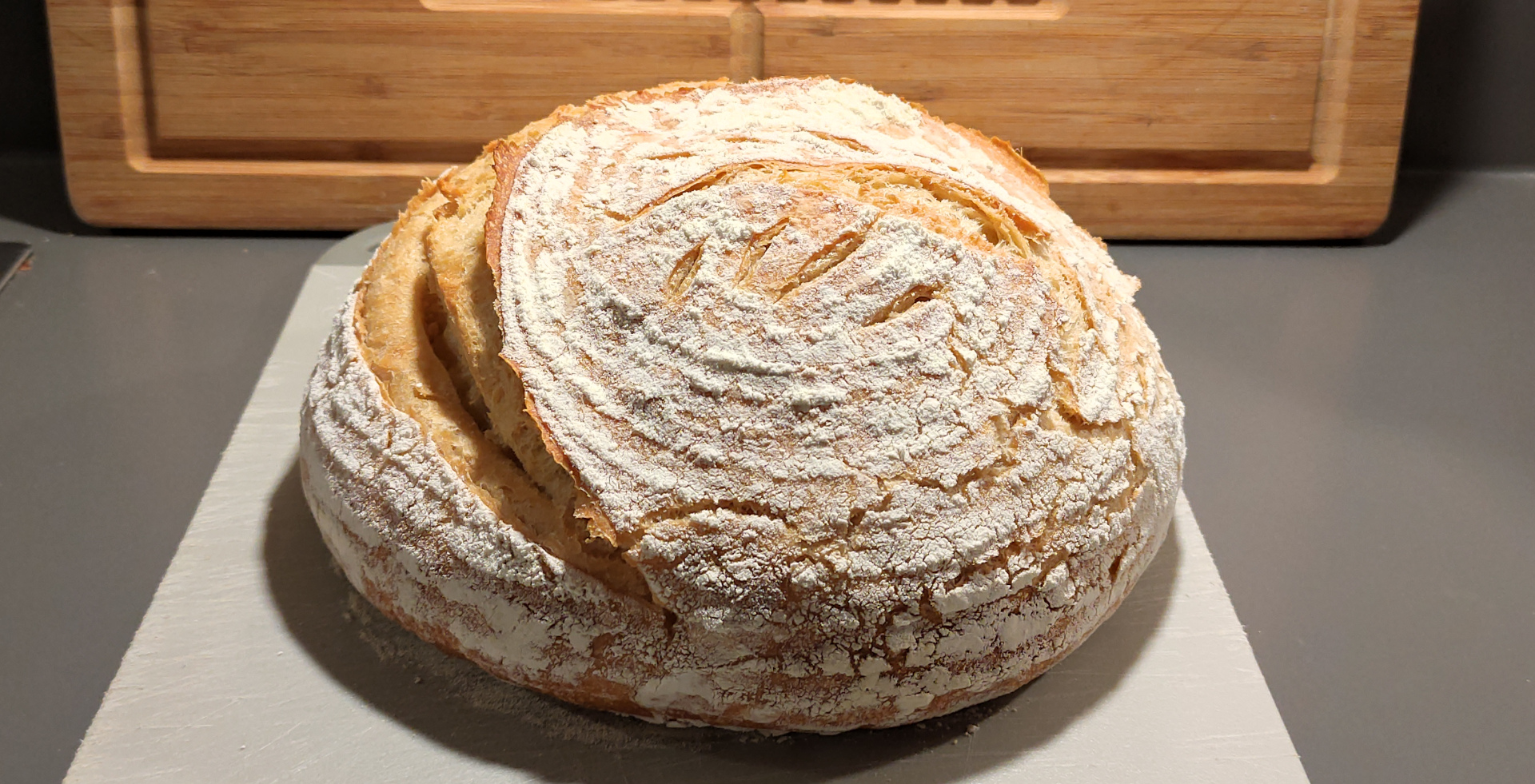 Ein goldbraunes Brot liegt auf einem Kunststoffbrettchen auf einer grauen ArbeitsflÃ¤che. Es ist an der linken Seite tief eingeschnitten und aufgegangen.