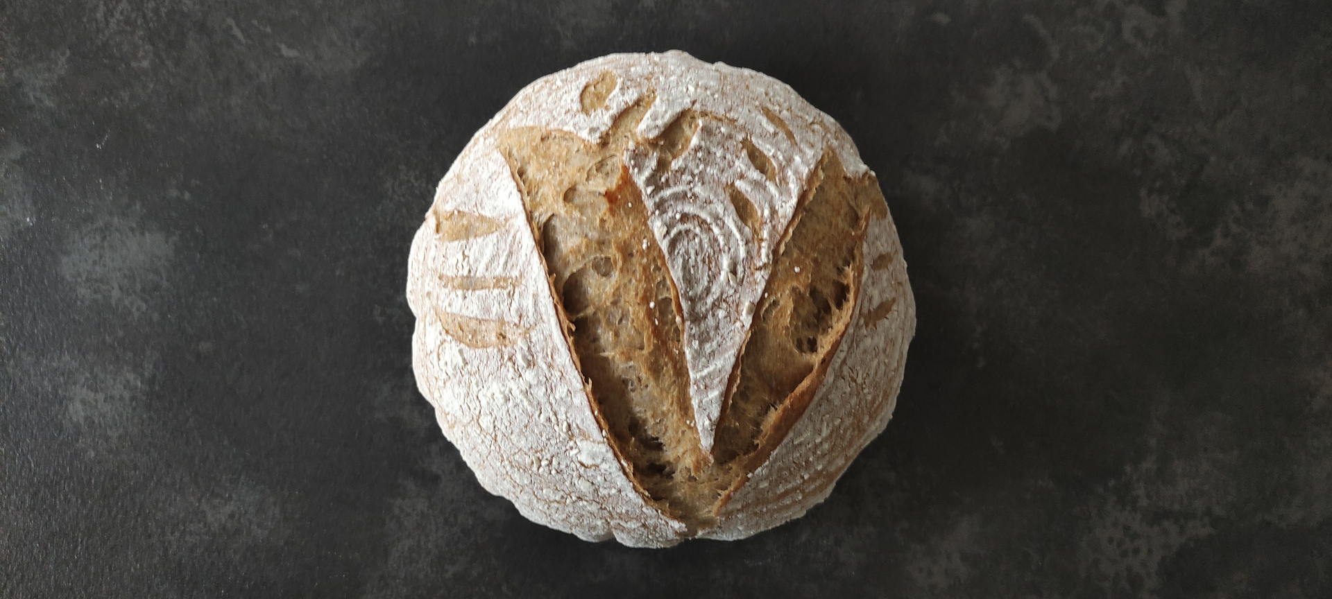 Fertig ausgebackenes Brot mit Einschnitten, die wie ein V zusammen laufen. Mehllinien verlaufen im Kreis rund Ã¼ber das gesamte Brot